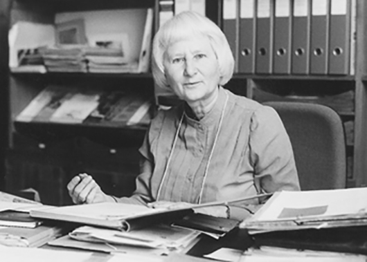 Dr. Gisela Reuther blättert in einem Buch und schaut in die Kamera