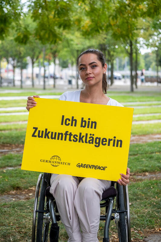 Eine Frau, die in einem Rollstuhl sitzt, hält ein gelbes Schild hoch, auf dem steht: Ich bin Zukunftsklägerin. Darunter die Logos von Germanwatch und Greenpeace.