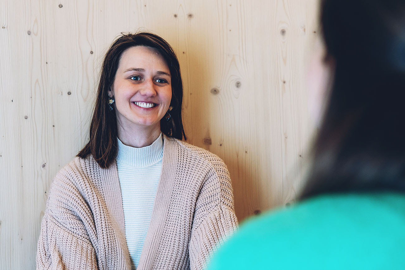 Frau mit rosa Strickjacke lächelt einer ihr gegenübersitzenden Person zu, im Hintergrund eine helle Holzwand.
