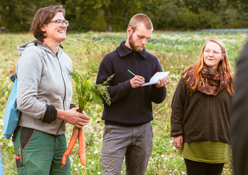 Eine junge Frau und ein junger Mann stehen auf einem Feld und hören einer Seminarleiterin zu, die zwei Karotten in der Hand hält und etwas erklärt.