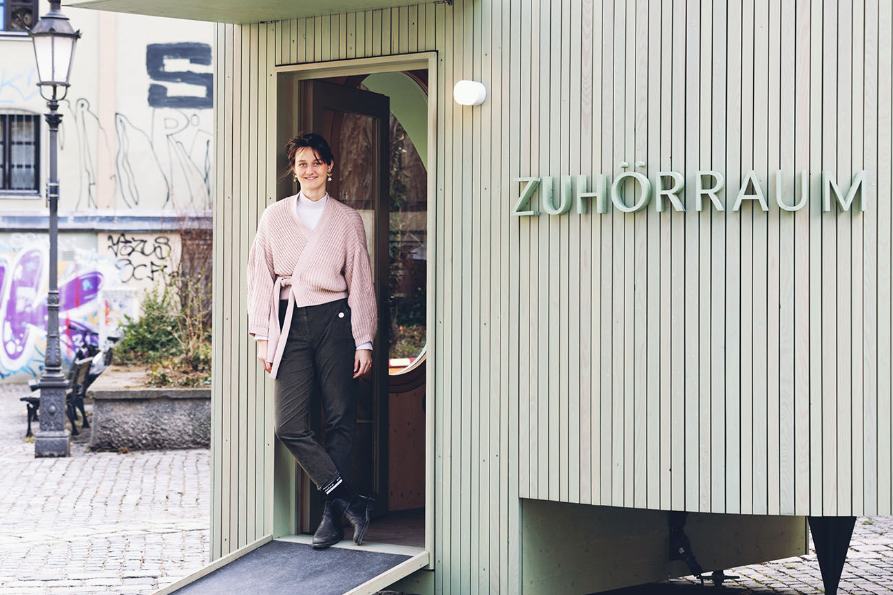 Lächelnde Frau in rosa Strickjacke steht in der offenen Tür eines mintgrünen Holzpavillons mit der Aufschrift „Zuhörraum" auf einem gepflasterten Platz.